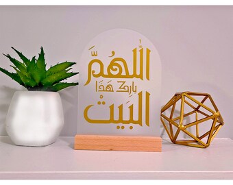 Op de afbeelding: Een wit acryl bord met een gouden Arabisch kalligrafie ontwerp. Het bord heeft de tekst "Allahumma innaka afuwwun tuhibbul afwa". Het bord staat op een houten standaard.