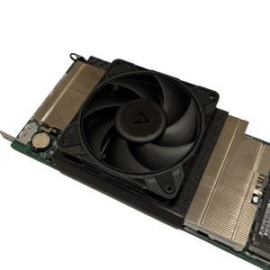 May include: A computer graphics card featuring a large black cooling fan with the Arctic logo in the center. The fan is mounted on a metallic heatsink with numerous thin cooling fins, attached to a green circuit board with a small silver battery.
