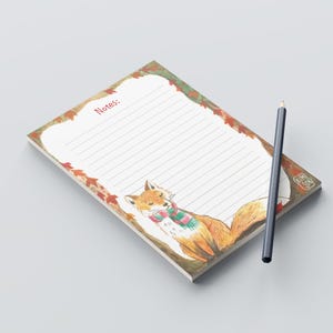 Könnte beinhalten: Ein Notizblock mit einem Fuchs, der einen Schal trägt, auf dem Cover. Der Notizblock hat einen weißen Hintergrund mit liniertem Papier und das Wort "Notizen:" in rot oben gedruckt. Der Fuchs ist von einem Rand aus Herbstblättern umgeben.