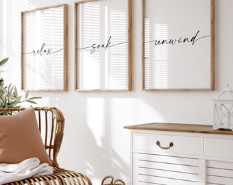 May include: Three framed prints with the words "relax", "soak", and "unwind" in a modern calligraphy style. The prints are hanging on a white wall above a white cabinet with a wooden top. A wicker chair with a brown cushion and a white throw blanket is in the foreground.