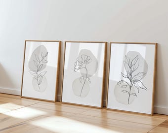 Puede incluir: Tres láminas de arte botánico enmarcadas. Cada lámina presenta un dibujo lineal minimalista de una planta o flor en negro sobre un fondo gris claro. Los marcos son de color marrón claro y la obra de arte se exhibe en una pared blanca.