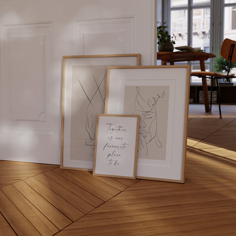 May include: Three framed prints with minimalist line art designs. The prints are in gold frames and are leaning against a white wall. The floor is made of wood planks. The text on one of the prints reads "Together is our favourite place to be."