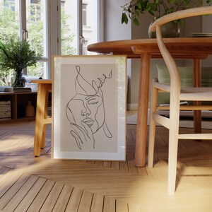 May include: A framed line art print of two faces, one overlapping the other, on a beige background. The print is hanging on a wall in a room with wooden furniture.
