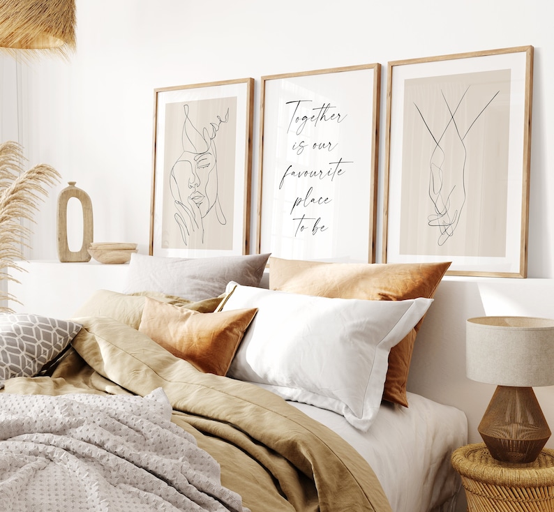 May include: Three minimalist line art prints in wooden frames on a beige wall above a bed with brown linen bedding. The prints feature a line drawing of a face, the text "Together is our favourite place to be", and a line drawing of two hands holding each other.