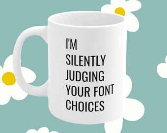 Könnte beinhalten: Eine weiße Kaffeetasse mit schwarzem Text, der "Ich beurteile deine Schriftarten stillschweigend" lautet, vor einem hellblauen Hintergrund mit weißen Gänseblümchen.
