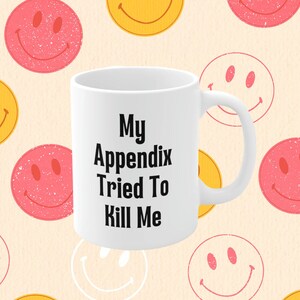 May include: White coffee mug with black text that reads "My Appendix Tried To Kill Me". The mug is surrounded by a pattern of yellow and pink smiley faces.