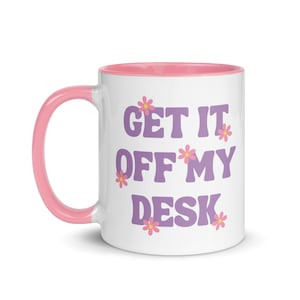May include: White ceramic mug with a pink rim and handle. The mug has a purple design that says "Get it off my desk" with pink flowers around the text.
