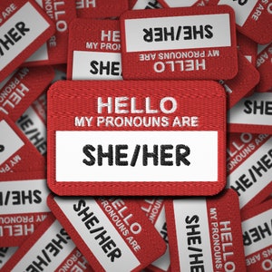 May include: Red fabric name tags with white text that reads "Hello, my pronouns are she/her".