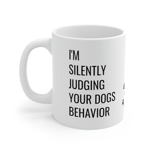 May include: White ceramic coffee mug with black text that reads "I'm silently judging your dogs behavior" with paw prints on the right side.
