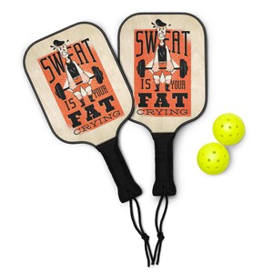 May include: Two pickleball paddles with black handles and the text "SWEAT IS YOUR FAT CRYING" on the paddle face. The paddles have a wooden appearance with a graphic of a weightlifter. Two yellow pickleballs are also in the image.