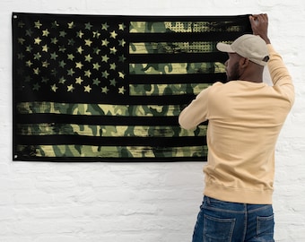 May include: A person hangs a black and green American flag with a camouflage pattern on a white brick wall. The flag has a distressed look and is made of fabric.