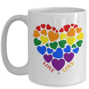 May include: White ceramic mug featuring a heart design composed of colorful hearts in rainbow colors. The words "LOVE IS LOVE" are printed in red and purple. The mug has a curved handle.
