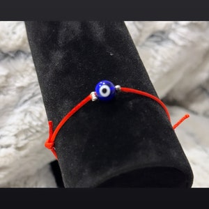 Puede incluir: Un brazalete de cuerda roja con un amuleto de ojo turco azul. El amuleto tiene un centro blanco.
