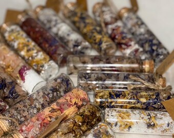 May include: A collection of glass tubes filled with colorful dried flowers and salts. Each tube has a cork stopper and a small brown tag. The contents vary in color, including pink, yellow, blue, and white, creating a visually appealing display.