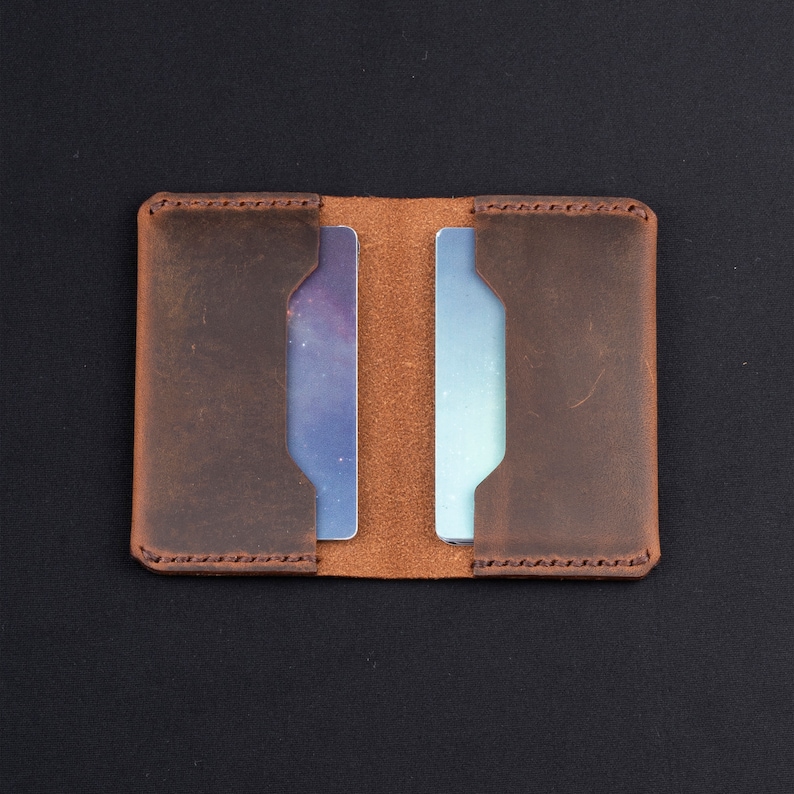 May include: A brown leather card holder with two slots for cards. The holder is open and the slots are empty.