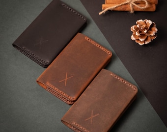May include: Three leather wallets in shades of brown, each with an embossed "X" design. The wallets are arranged on a dark surface, with a pine cone and cinnamon sticks in the background. The wallets have stitched edges.