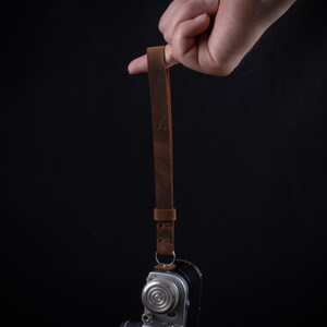 May include: A brown leather camera strap with a stitched design and an 'X' embossed on the strap. The strap is attached to a silver camera.