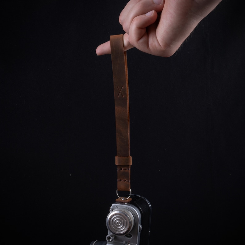 May include: A brown leather camera strap with a stitched design and an 'X' embossed on the strap. The strap is attached to a silver camera.