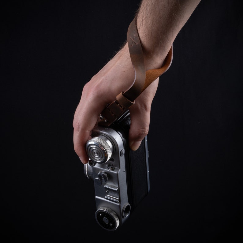 May include: A silver camera with a brown leather strap attached to the wrist. The camera has a black lens and a silver dial on the side.