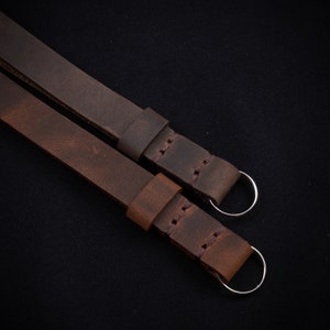 May include: Two brown leather camera straps with silver metal rings. The straps are stitched together and have a distressed look.