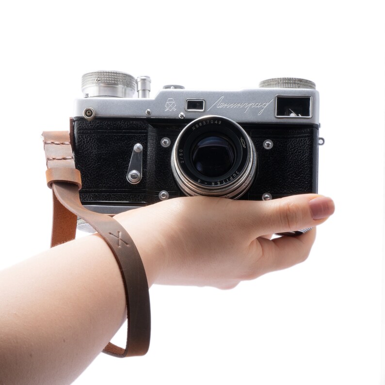May include: A silver and black vintage camera with a leather strap. The camera has a lens with the text "Jupiter-8" on it. The camera is being held by a hand.