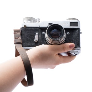 May include: A silver and black vintage film camera with a leather strap. The camera has a lens with the text "Jupiter-8" printed on it.