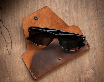 May include: A brown leather glasses case with a flap closure. The case is open and a pair of black sunglasses are inside.