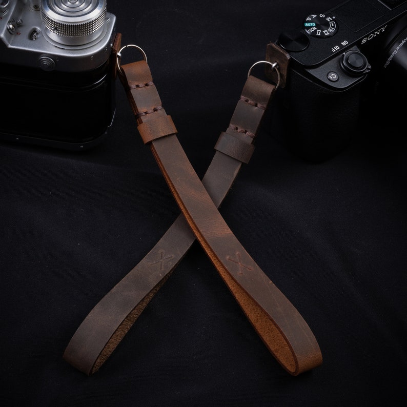 May include: Two brown leather camera straps with silver metal rings and a cross-shaped design. The straps are crossed over each other.