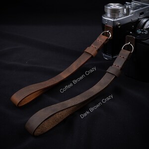May include: Two leather camera straps in different shades of brown. The strap on the left is labeled "Coffee Brown Crazy" and the strap on the right is labeled "Dark Brown Crazy".