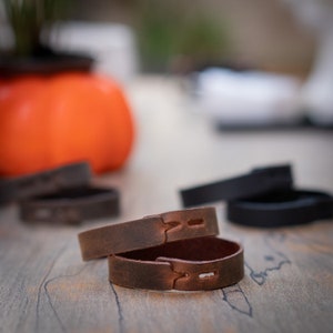 May include: Two brown leather bracelets with stitching detail. The bracelets are stacked on top of each other.