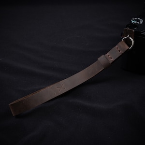 May include: A brown leather camera strap with a stitched edge and a small 'X' embossed on the strap.