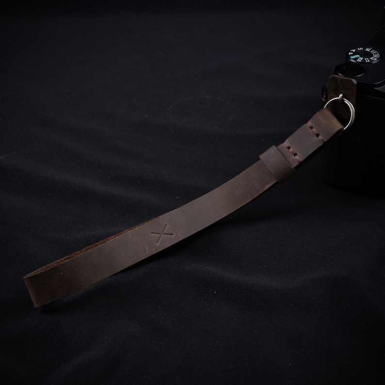 May include: A brown leather camera strap with a stitched edge and a small 'X' embossed on the strap.