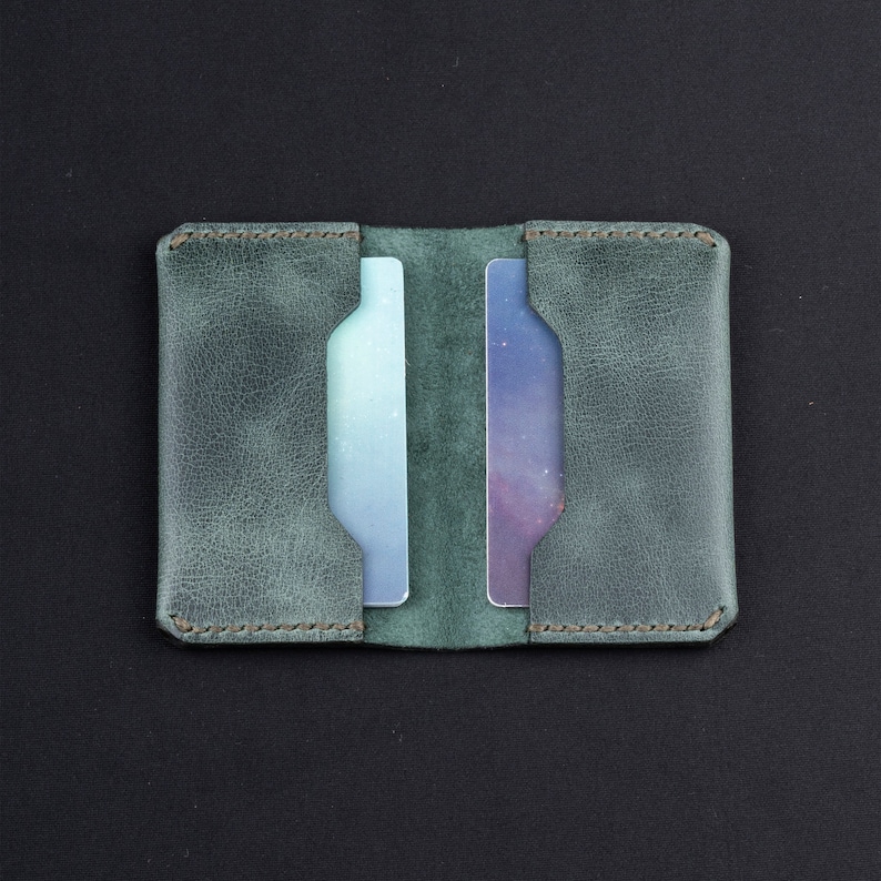 May include: A green leather card holder with two slots for cards. The holder has a distressed look and is stitched around the edges.