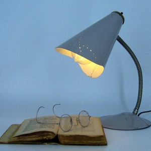 May include: A grey metal desk lamp with a cone-shaped shade featuring a pattern of small holes. The lamp has a flexible arm and a round base. The lamp is turned on and illuminating a book and glasses.