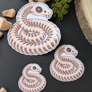 Könnte beinhalten: Drei gestanzte Aufkleber mit der Illustration eines zusammengerollten Schlangenskeletts. Jede Schlange hat eine cremefarbene Knochenstruktur, ein dunkelbraunes Inneres und einen Schädel mit einem kleinen weißen Herz in der Augenhöhle.