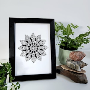 May include: A black and white mandala design printed on white paper and framed in a black frame. The frame is sitting on a white surface.