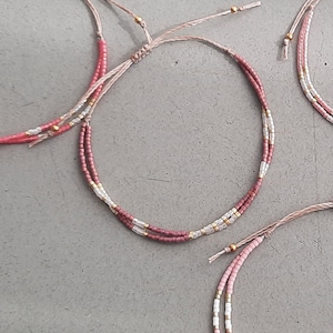 May include: Two delicate bracelets featuring pink, white, and gold beads. The bracelets are tied with a simple knot and are made with a thin, light brown cord.