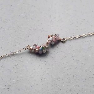 Op de afbeelding: Een delicate gouden ketting met een cluster van kleurrijke edelstenen in een traanvorm. De stenen zijn roze, groen en wit.