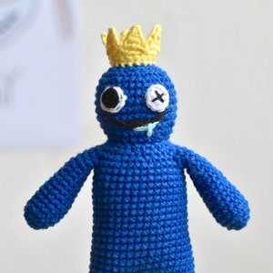 Puede incluir: Un juguete de crochet azul con una corona amarilla, un ojo negro y un ojo blanco con una X. El juguete tiene una sonrisa torcida y una baba verde.