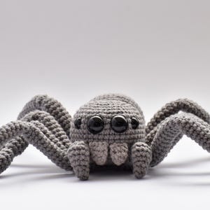 Puede incluir: Una araña de ganchillo gris con un cuerpo redondeado y ocho patas articuladas. La araña tiene ojos grandes y negros y una apariencia detallada y texturizada, lo que sugiere un artículo artesanal hecho a mano.