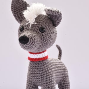 Puede incluir: Un perro de ganchillo gris hecho a mano con un mechón de pelo blanco y un collar a rayas rojas y blancas. El perro tiene ojos negros y una nariz negra. El perro está de pie sobre cuatro patas y tiene una cola pequeña.