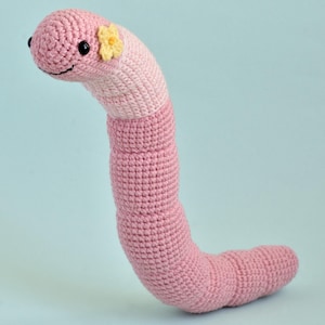 Peut inclure: Ver de terre rose en crochet, avec une tête rose clair et une fleur jaune. Le ver a des yeux noirs et une bouche souriante. Le fond est bleu clair.
