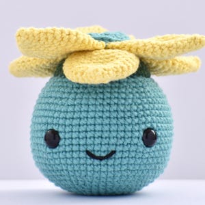 Puede incluir: Un juguete de peluche tejido a ganchillo con forma de flor sonriente. El cuerpo es turquesa, con ojos negros y una sonrisa cosida. Los pétalos de la flor son amarillos y verdes.
