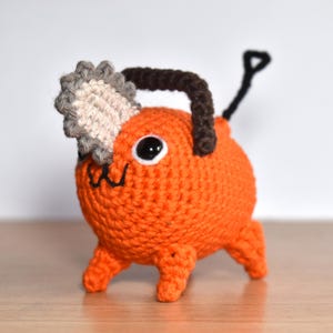 Könnte beinhalten: Ein orangefarbenes Häkeltier mit einem Kettensägenkopf und einem dunkelbraunen Griff. Das Spielzeug hat schwarze Augen und einen kleinen schwarzen Mund. Der Kettensägenkopf ist grau und beige. Es steht auf vier kleinen Beinen.