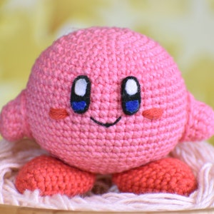 Puede incluir: Juguete de peluche rosa tejido a crochet con una cara sonriente, ojos azules y mejillas sonrosadas. El juguete está sentado sobre un fondo de hilo blanco.