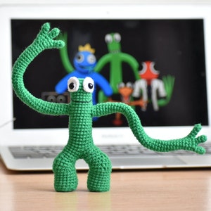 Puede incluir: Una figura verde hecha a mano con ganchillo, con grandes ojos blancos y brazos extendidos. La figura está frente a una pantalla de ordenador portátil que muestra personajes de dibujos animados en azul, verde y naranja. La figura está sobre una superficie de madera.