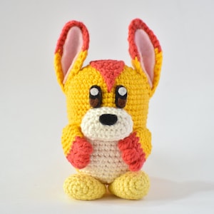 May include: A handmade crochet dog toy in yellow, red, and white. The dog has large ears with pink inner ears, brown eyes, and a white muzzle. The toy is standing on two yellow feet.