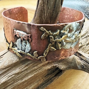 May include: A copper cuff bracelet with an embossed design. The bracelet features a figure, leaves, and a floral pattern in silver and gold tones. The cuff is approximately 2 inches wide.