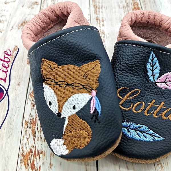 May include: A pair of black leather baby shoes with embroidered designs. One shoe features a fox with a feather headdress and the other has leaves and the name "Lotta".