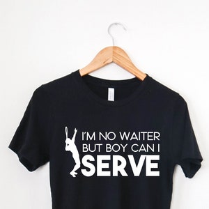 May include: Black t-shirt with a white graphic of a tennis player serving a ball. The text on the shirt reads "I'm no waiter but boy can I serve."